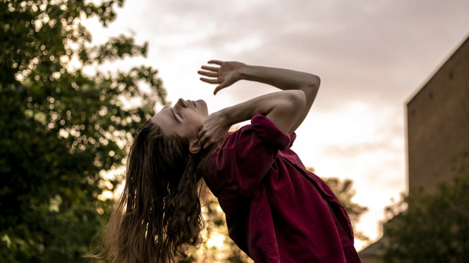 Young male dancer with long blond hair wearing a burgundy t-shirt stretches backwards. He stands in a leafy setting outdoors beneath a pink sunset.