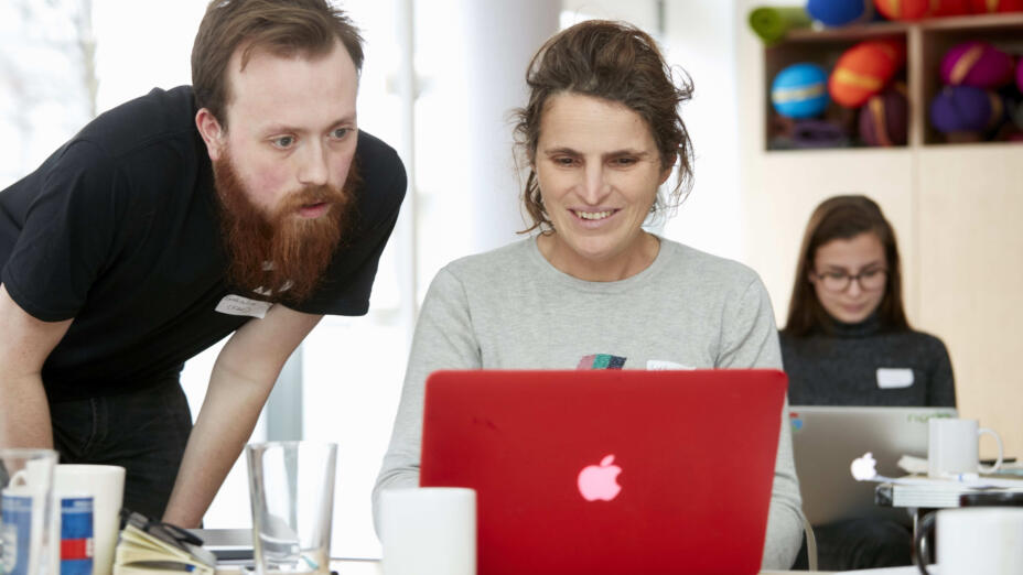 Woman wearing a grey shirt sits in front of red mac latop and smiles while a young man with a beard and dressed in black helps her out. They look at the screen together and collaborate.