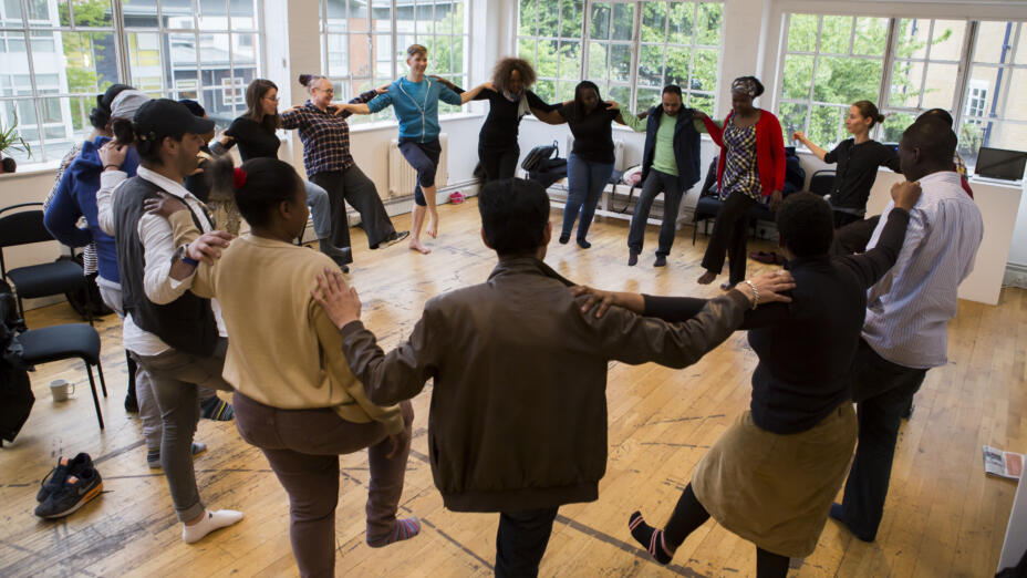 A large group of around 20 people are in a bright room in a circle. They're each holding onto the shoulders of the person either side of them, with one foot lifted off the ground in a game