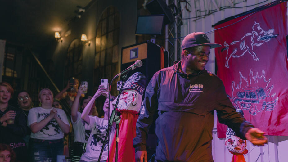 A young man is on stage wearing a Museum of Homelessness rain jacket. He is in front of a microphone with a dozen or so audience members photographing and cheering.
