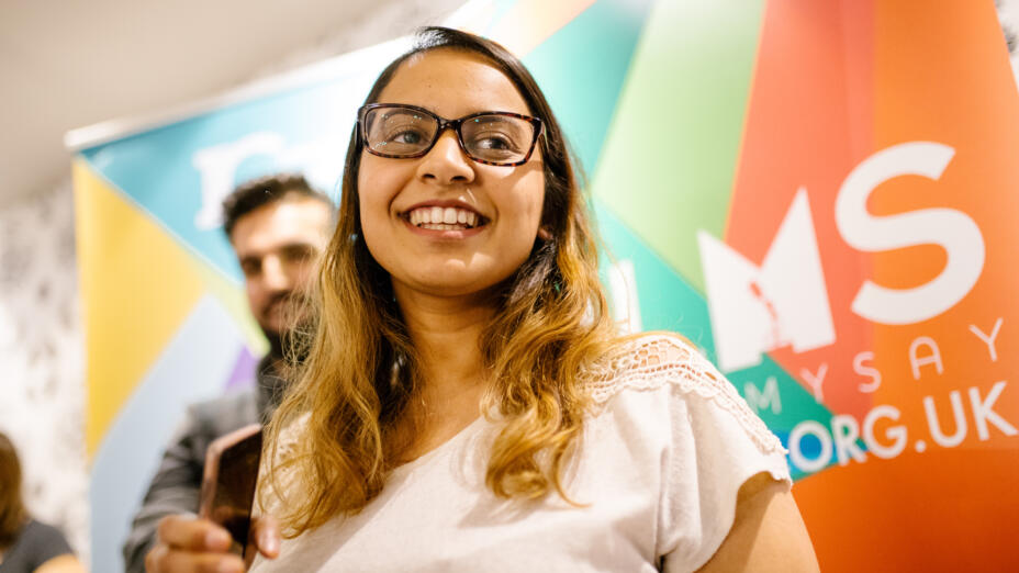 Young woman wearing glasses and a beige top stands smiling in front of a brightly coloured banner for My Life My Say
