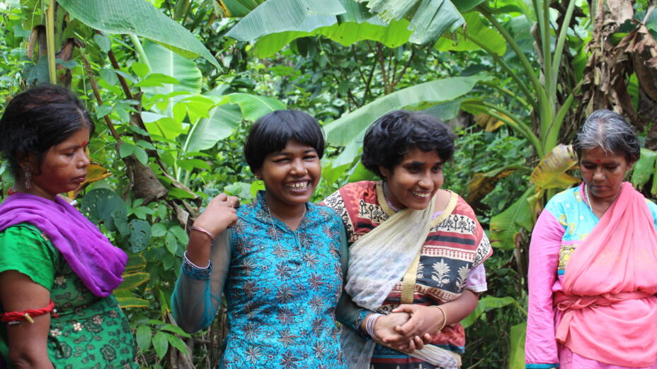 Four women in brightly coloured outfits from Iswar Sankalpa's Nayagram assisted community living programme stand together outside in front of a lush green background.