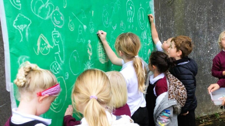 A group of school children draw smiley faces and doodles on a large piece of green paper taped to a wall.