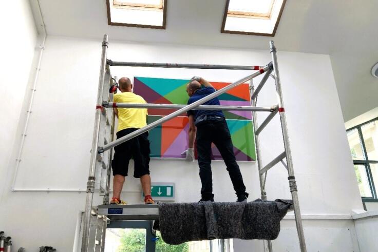 Two technicians on scaffolding install part of a large abstract artwork by Richard Bell in Newlyn Primary School.