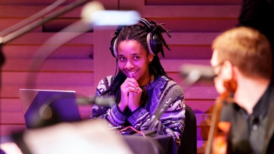 Composer Nneka Cummins sits in front of a laptop with headphones on while listening to a fellow musician who is holding a violin.