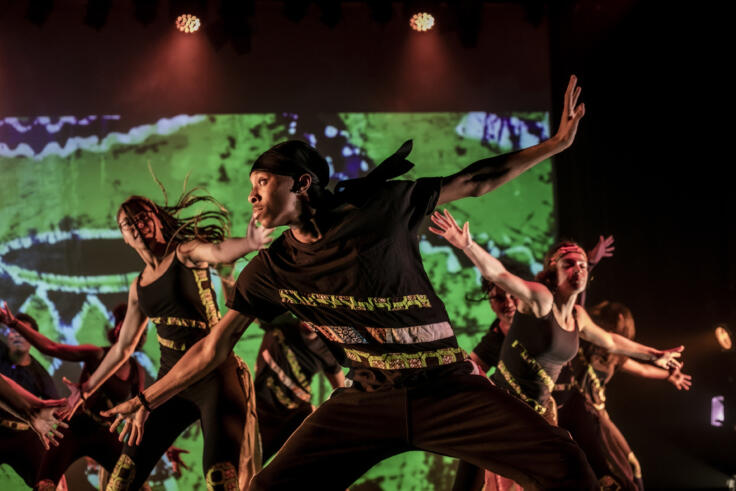 Young dancers are performing on a stage with a vibrant green background and two red spotlights over them