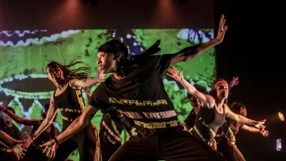 Young dancers are performing on a stage with a vibrant green background and two red spotlights over them