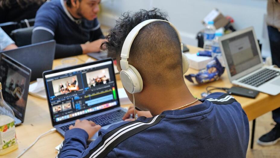 Two young men sit at laptops wearing headphones and using video editing software.