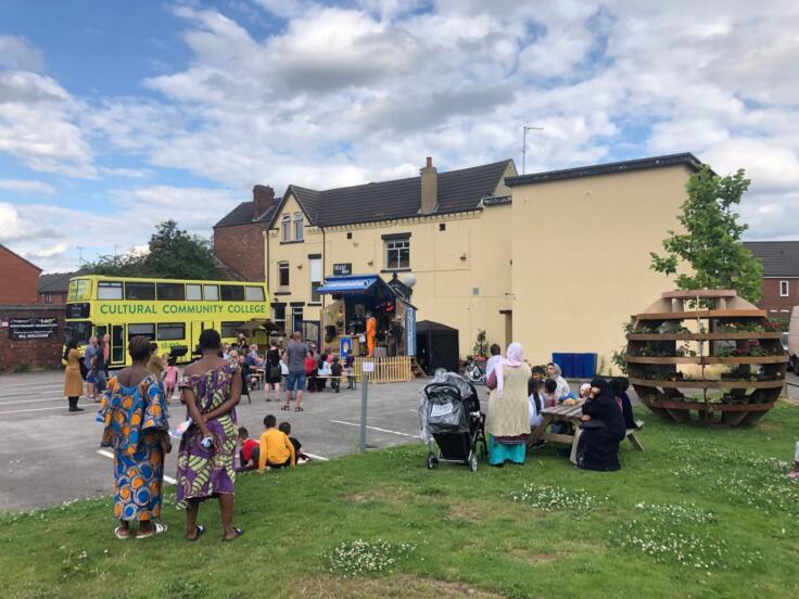 Yellow and green bus that says 'Cultural Community College'. A group of people attend an outdoor performance in front of a large cream stucco building. There's a wooden spherical sculpture on the right.