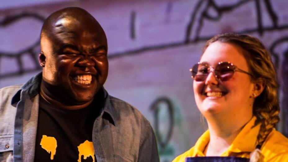 Two actors are standing next to each other, both with big smiles. The man on the left is wearing a black t-shirt with a big yellow graphic of a dripping smile, and the woman on the right is wearing a bright yellow shirt with blue dungarees, with a pin badge of a big smile similar to the man on the left's shirt