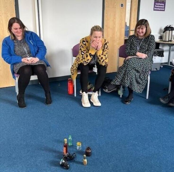 Three teachers sit next to one another on chairs in a blue-carpeted room. They're smiling as look at a collection of bottles in the middle of the floor in front of them,