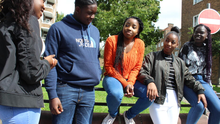 A group of young people are outside a block of flats, sitting lined up on a wall nearby and speaking to each other