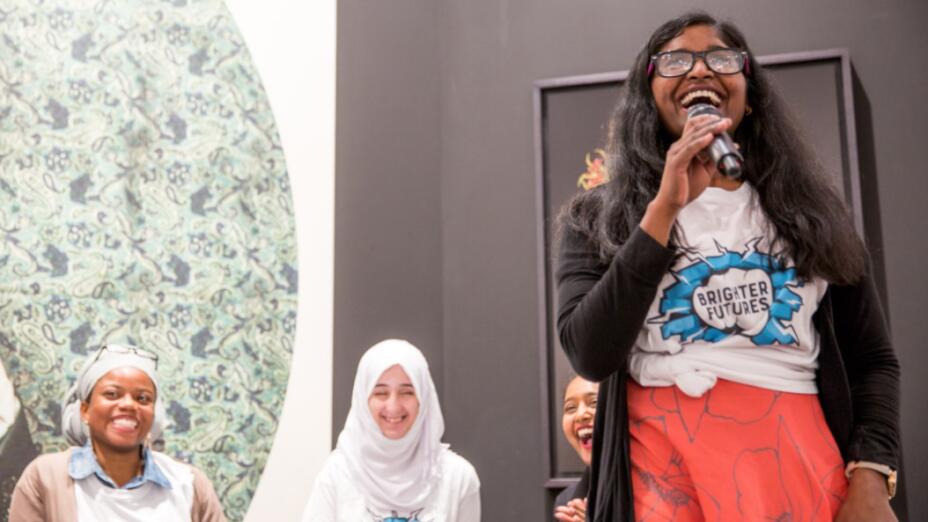 Four young women are on a stage, all smiling and laughing. A young girl is stood up in front, holding a microphone and smiling, wearing a t-shirt that says brighter futures