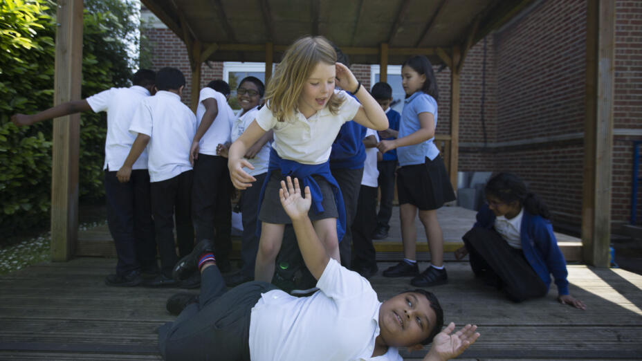 A group of pupils are standing on a deck outside and taking part in a drama-based session. A girl with blond hair stands over a young boy who is on the ground as they act out a scene happily together.