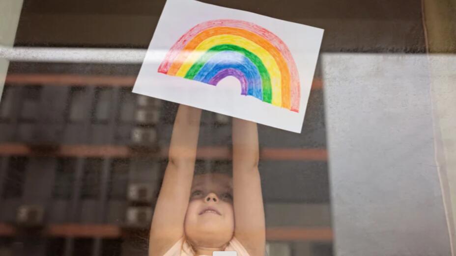Little girl holds a drawing of rainbow up to a window and you can see a block of flats in the reflection.