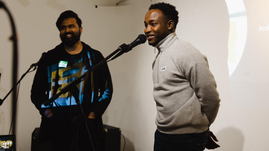 Two young men stand on stage smiling and speaking into microphones. One is wearing a brightly coloured t-shirt and a black hoodie and the other is wearing a grey cardigan and black trousers.