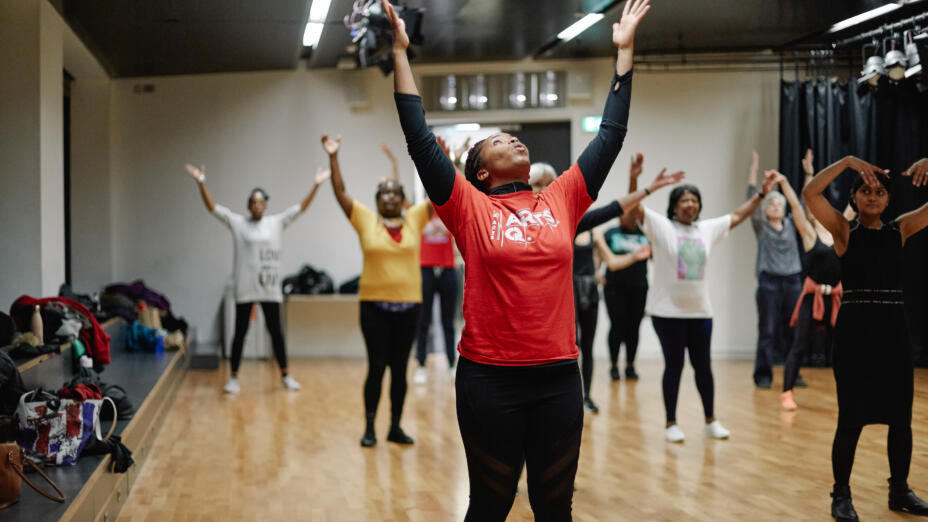 Group of dancers in bright t-shirts and black leggings raise their hands up together as they practice a dance in a rehearsal space with a wooden floor.