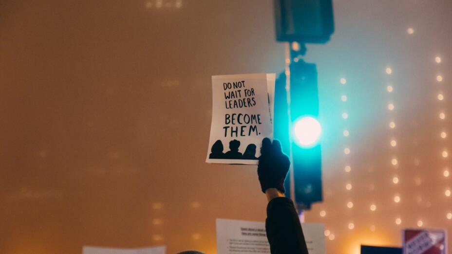Young person holds up a small black and white poster that reads: Don't wait for leaders, become them.