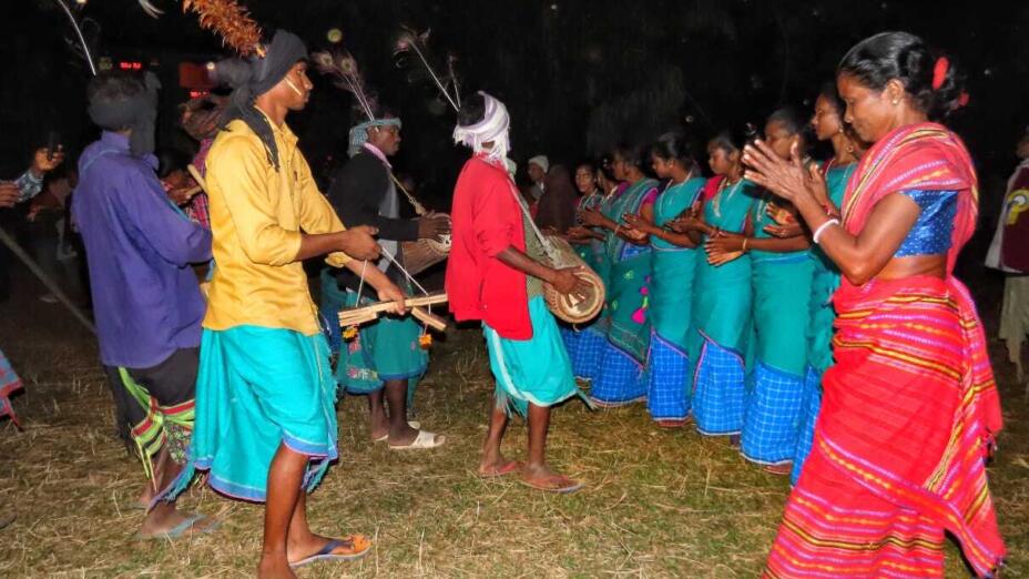 Woman in a red sari claps standing next to a line of women in turquoise saris.