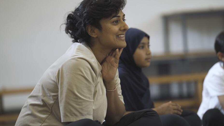 Teacher sits on the floor and holds her hand to chin smiling while leading children in an arts-based learning session.