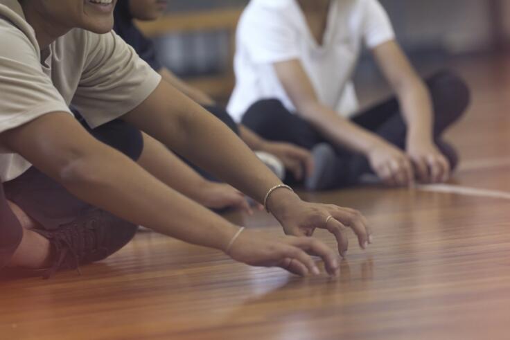 Three children sit cross-legged on the floor holding their hands in front of them and tapping their fingers on a wooden floor.