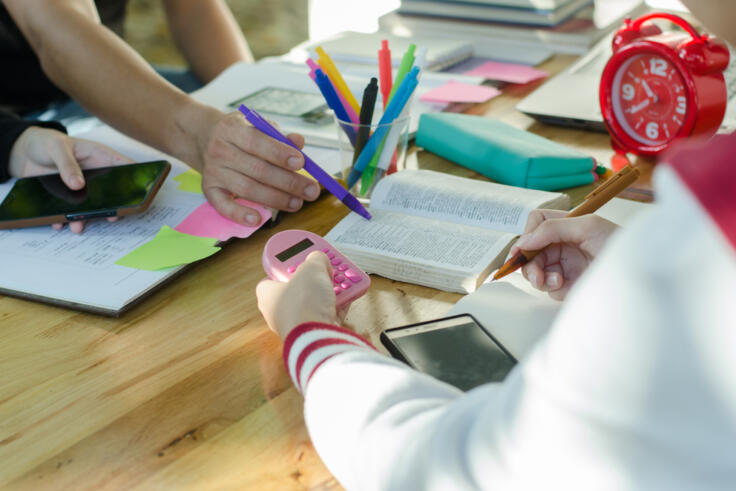 An image of 3 young women's hands as they work together at a table. They're holding calculators and there are books and post-its spread across the table.
