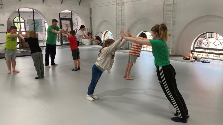 Pupils and artist practioners stand in pairs with their arms raised to the ceiling and leaning on each other's joined hands. They are all standing in a loft rehearsal space with concrete floors and white walls with arched windows.