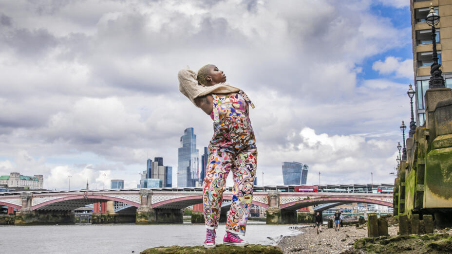 A young dancer wearing brightly patterned dungarees is posing on a river bank of the Thames in central London. Blackfriars bridge and a London skyline of skyscrapers is visible behind her, while she curls one arm behind her head and is turned to her side