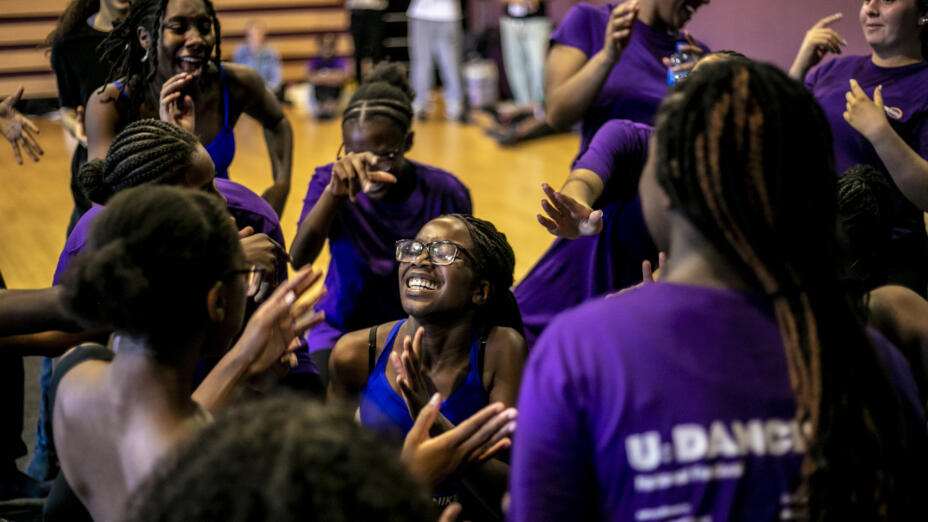 A group of young girls are standing in an informal circle and laughing. They are in a dance studio, with most wearing purple t-shirts
