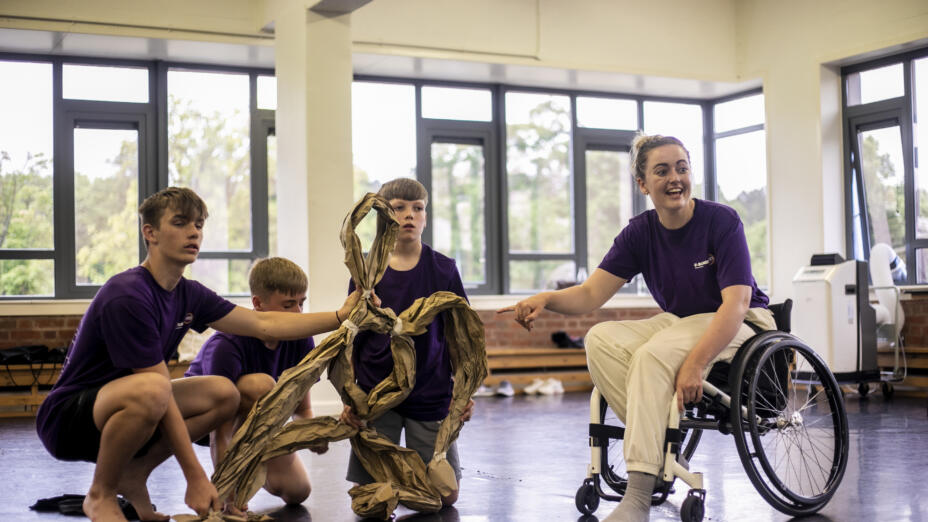Four young people are in a dance studio. Three young boys are holding a large abstract object crafted out of brown paper, while a young woman next to them in a wheelchair gestures to the object and smiles