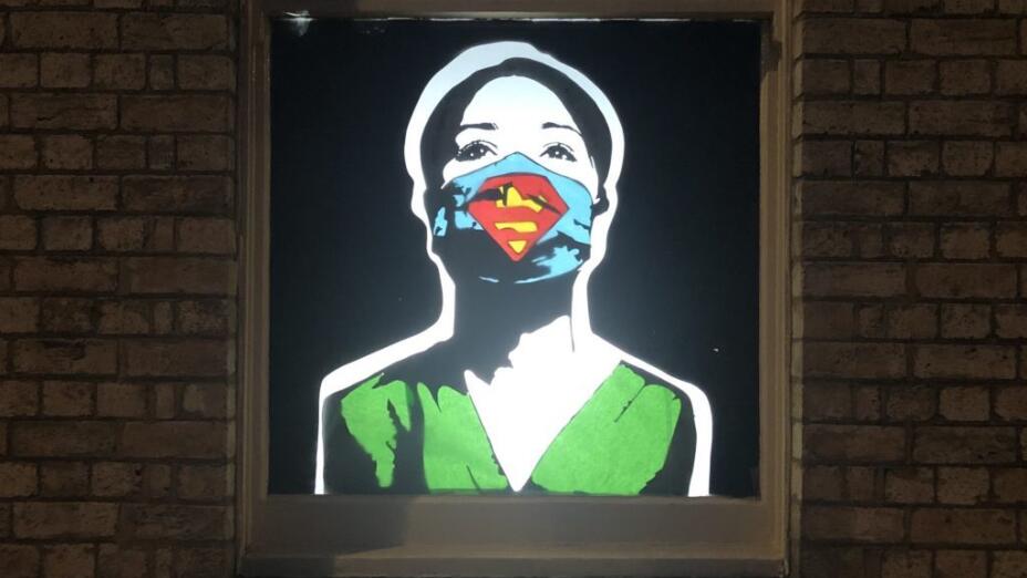 Graphic on a window of an NHS nurse in bright green scrubs and wearing a blue face mask with the Superman logo.