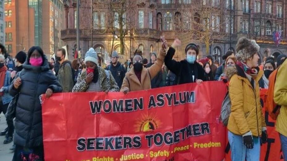 Campaigners from WAST Manchester hold a red banner that reads: Women Asylum Seekers Together. Human Rights. Justice. Safety. Peace. Freedom.