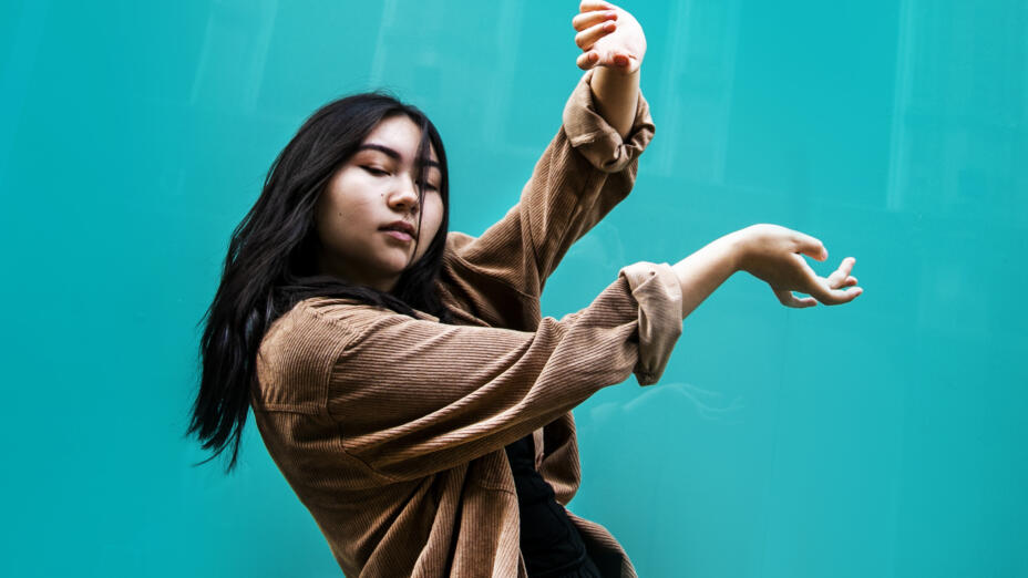 A young dancer is posed against a turquoise background. She is wearing a loose brown shirt, with sleeves rolled up, and has her hands raised out to her side