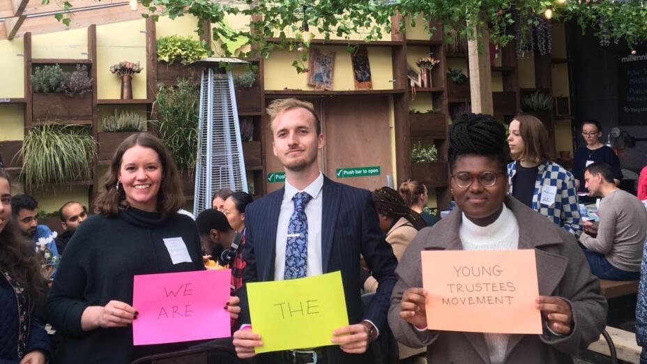 Three young people in work clothes stand against a leafy background holding signs on brightly coloured paper that say We are the Young Trustees Movement.